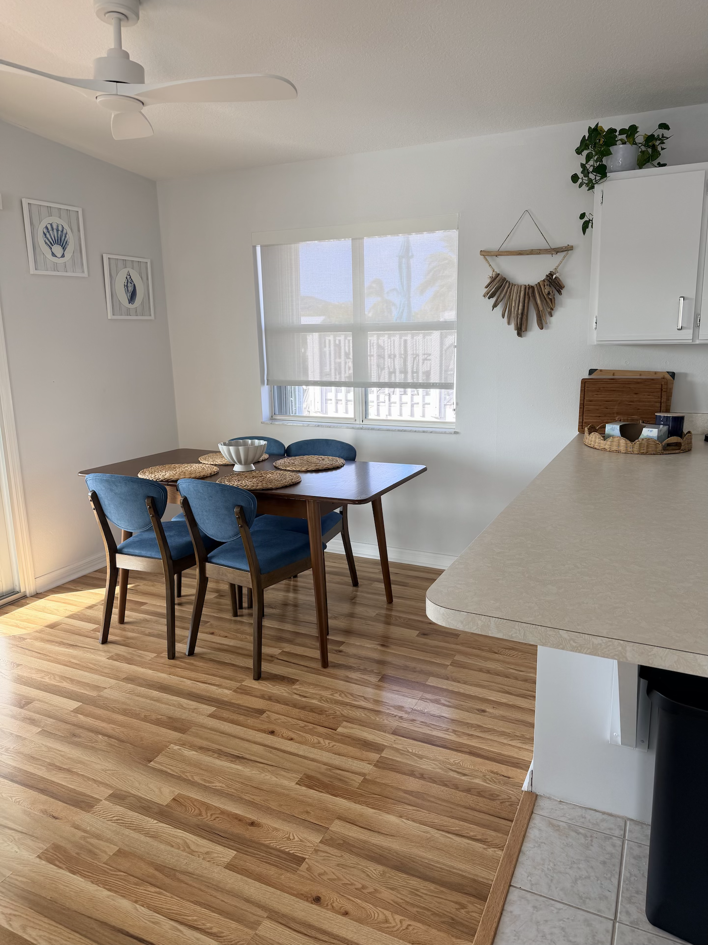 A dining area with a wooden table and four blue chairs, a decorative bowl, and a window with a light curtain, in a home with light-colored walls and wood flooring.