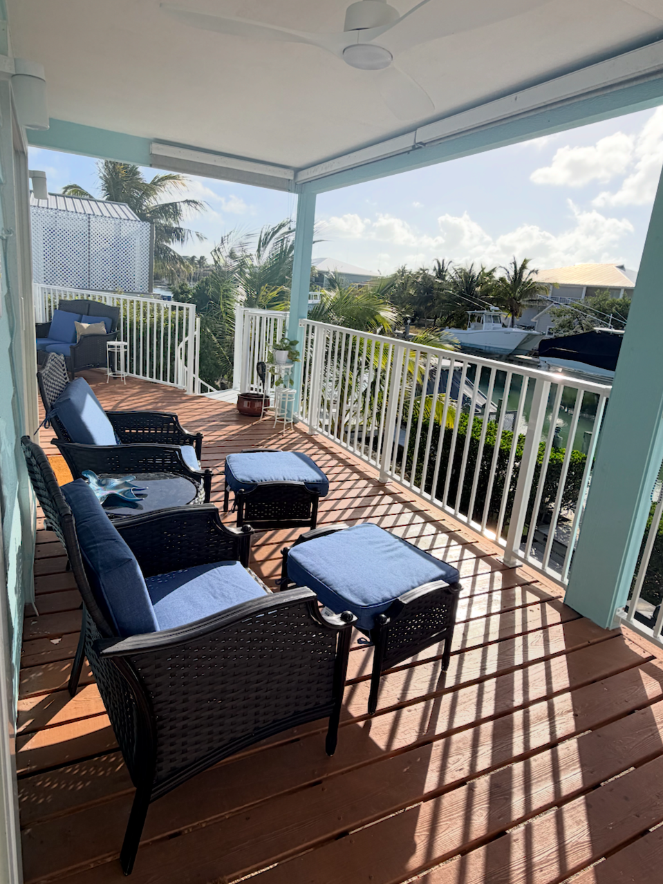 A screened porch overlooking a marina with boats, several black wicker chairs with blue cushions, and potted plants, under a ceiling fan with partly cloudy skies.