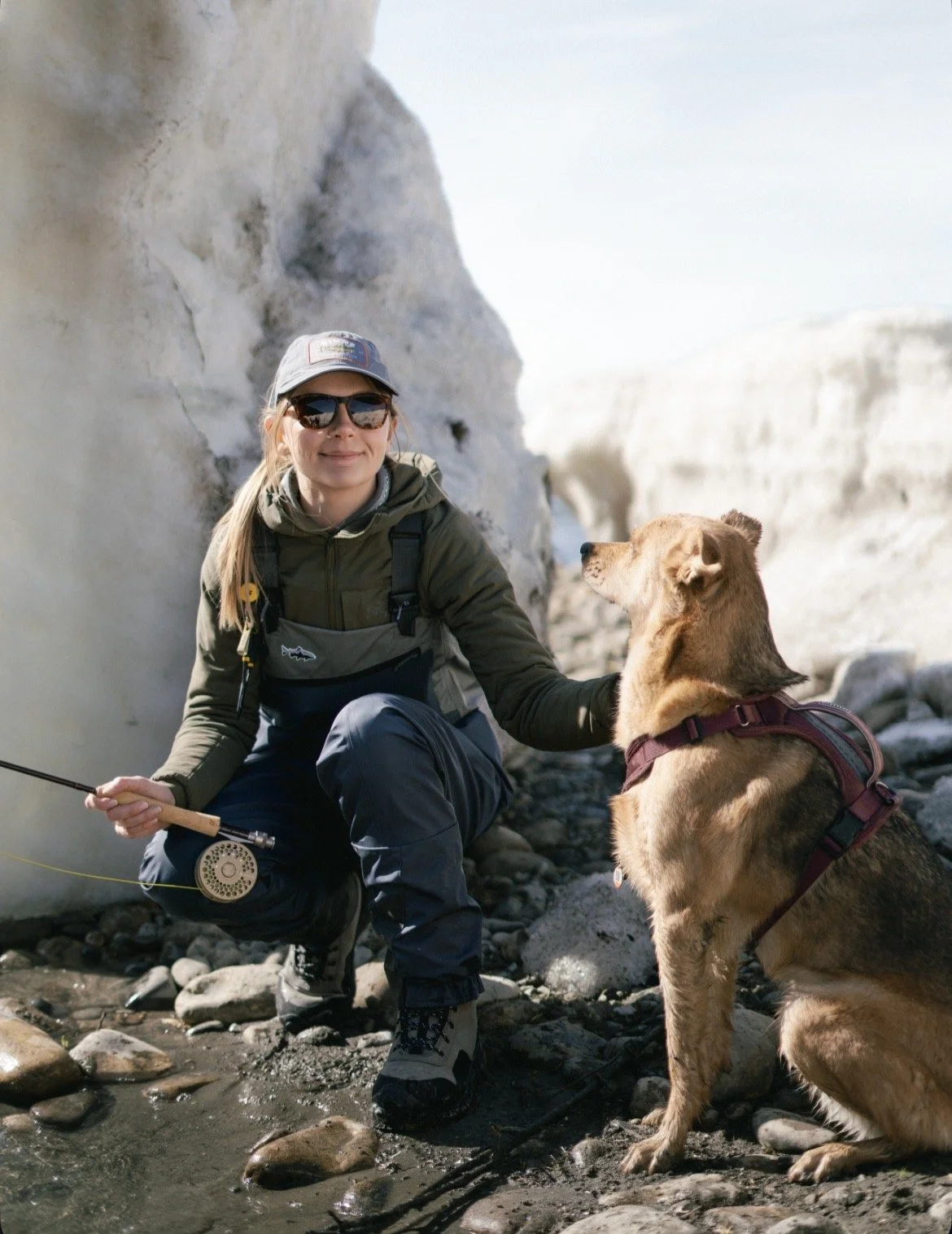 girl in waders holding fishing rod with her dog sitting next to her