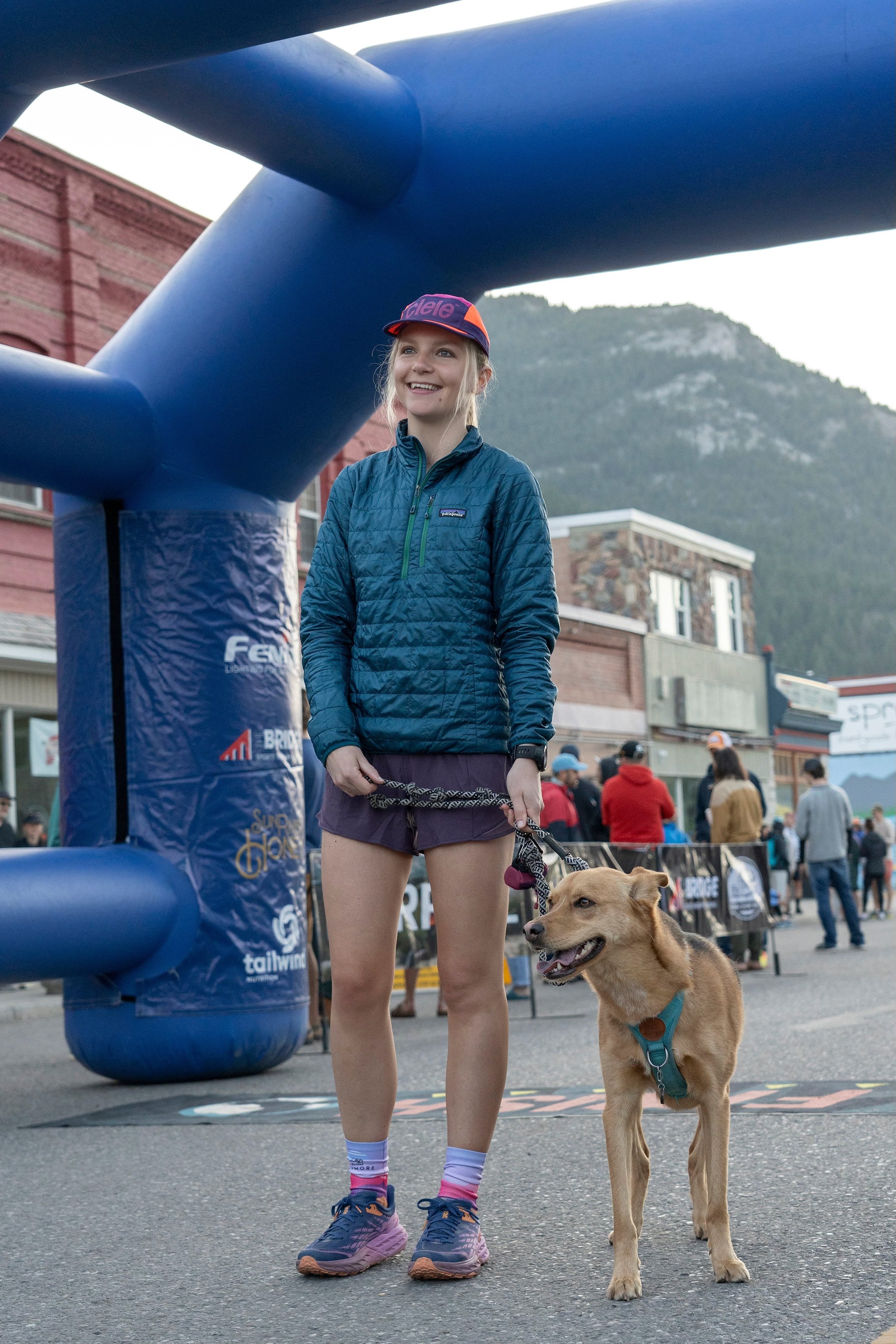 A woman in athletic attire with a dog under an inflatable arch at an outdoor race event, with mountains in the background and a crowd of people.