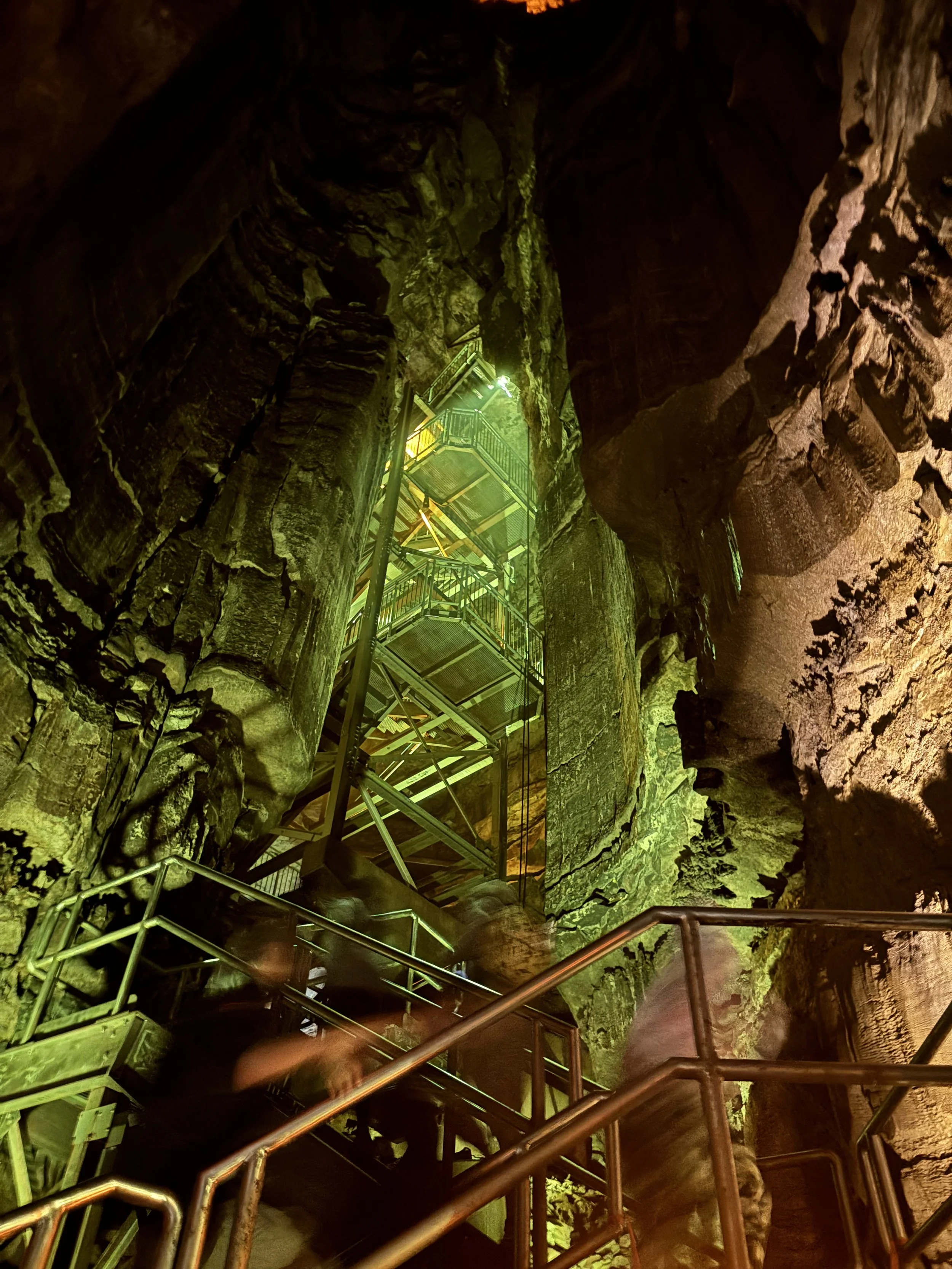 Inside a large cave with tall rock formations, a metal staircase with handrails leading upward, illuminated by greenish and yellow lighting.