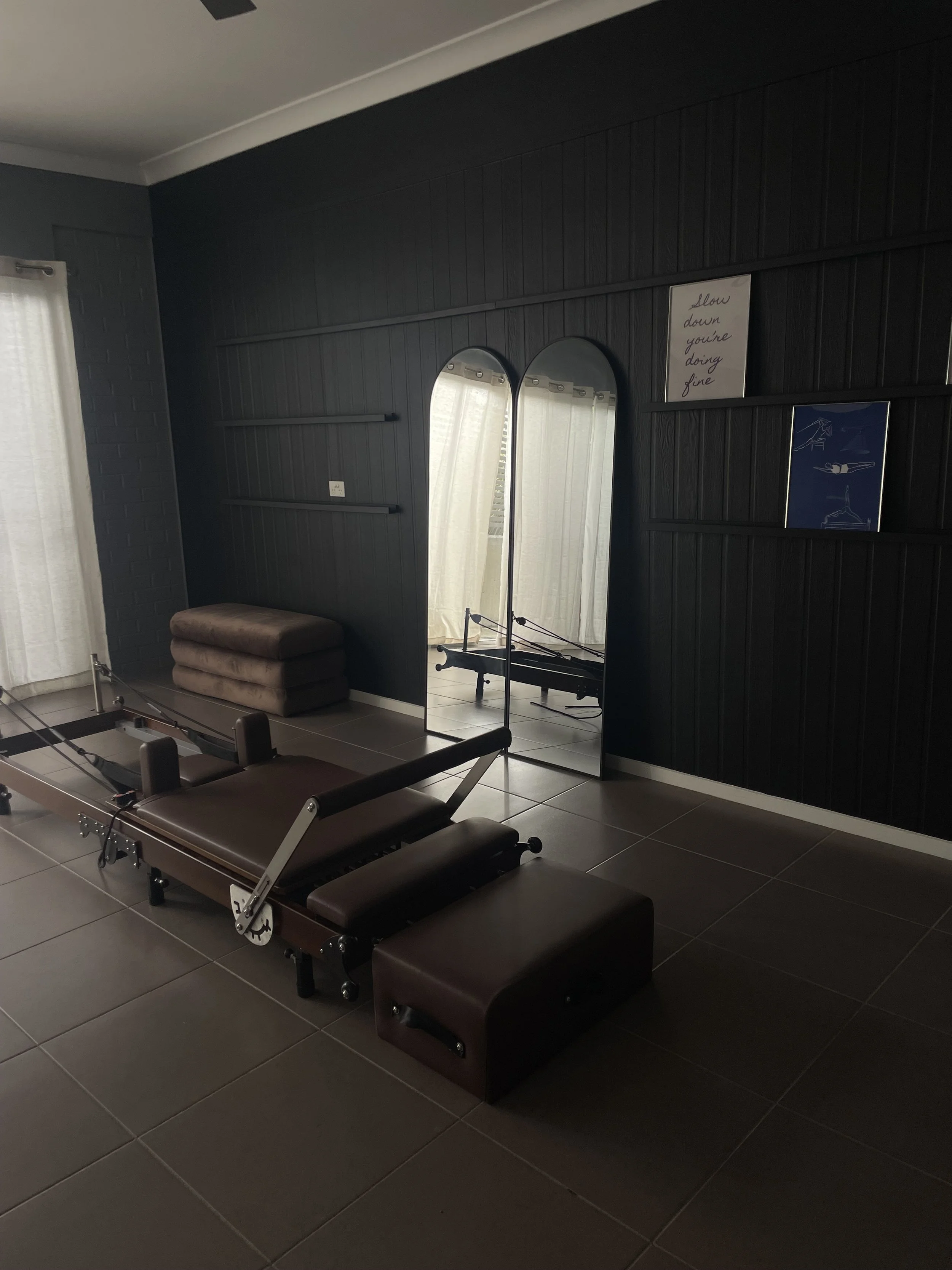 A Pilates reformer machine with brown padding in a dark room with black wood-paneled walls, a tall arched mirror, a bench, and inspirational art on the wall.