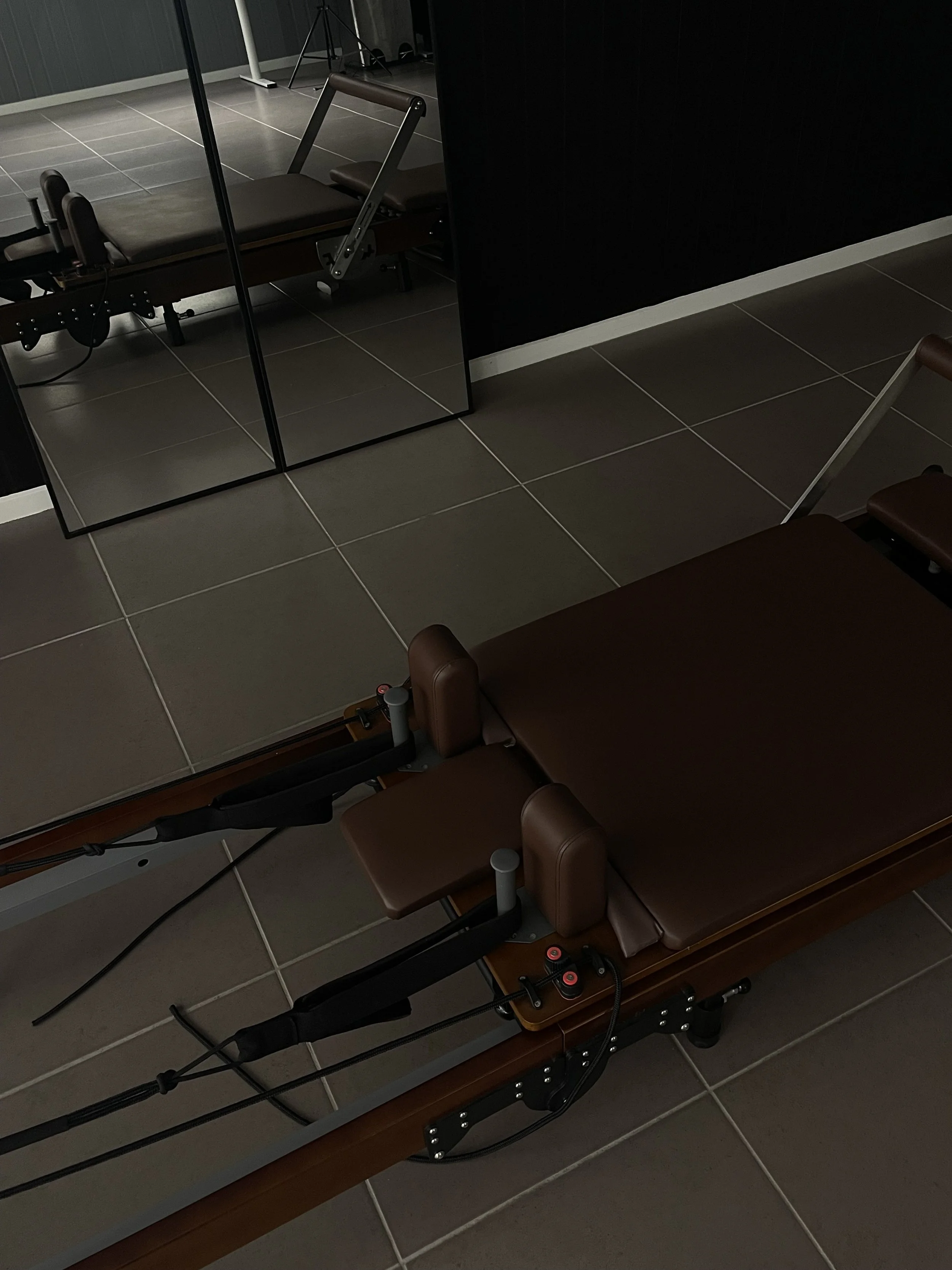 A brown reformer, positioned in a room with tiled floors and a mirror reflecting part of the room.