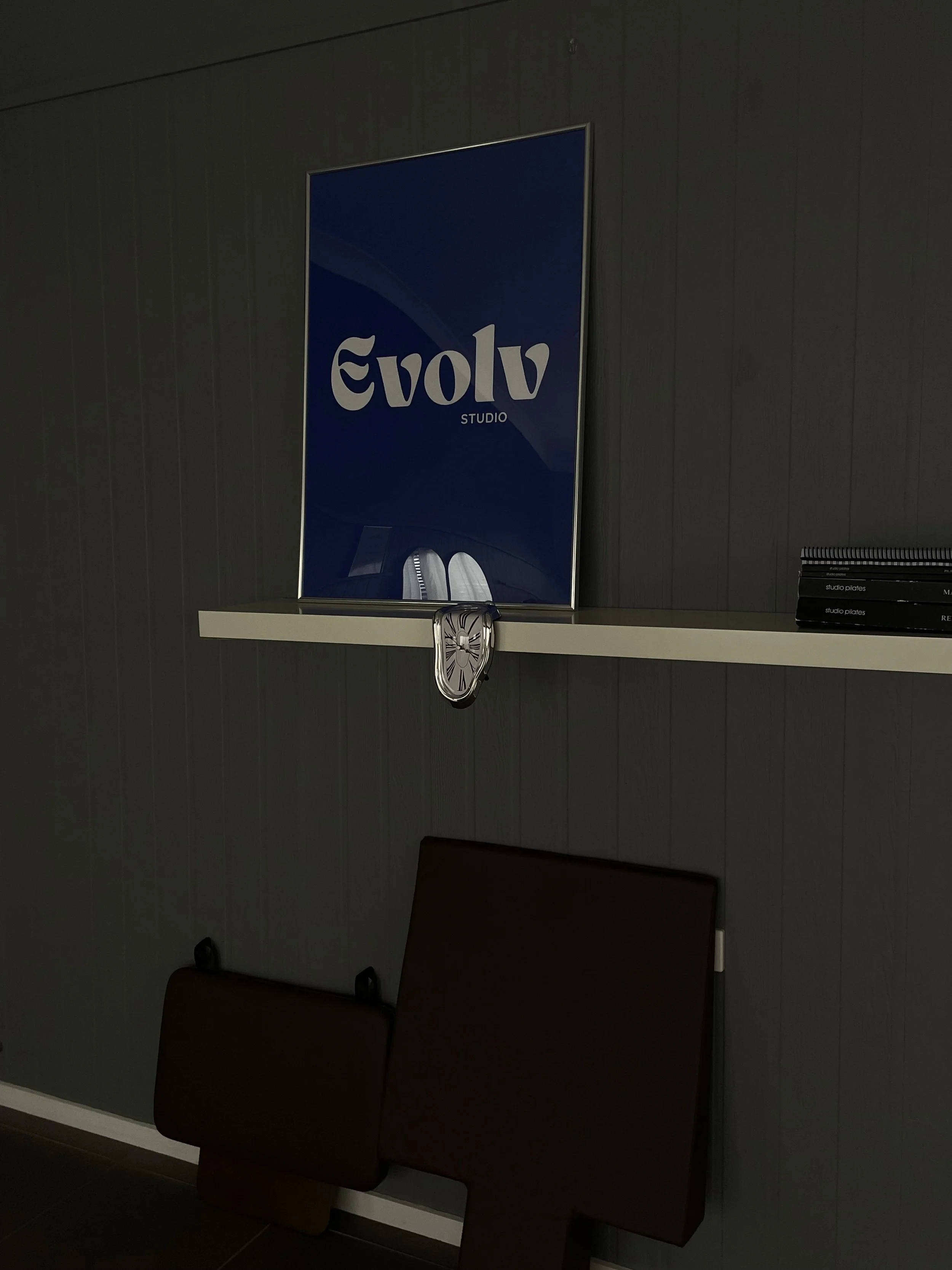 Interior of a room with grey wall paneling, a white shelf with a mirror clock hanging from it, and a large blue sign with "Evolv Studio" on it. Reformer equipment visible at the bottom.