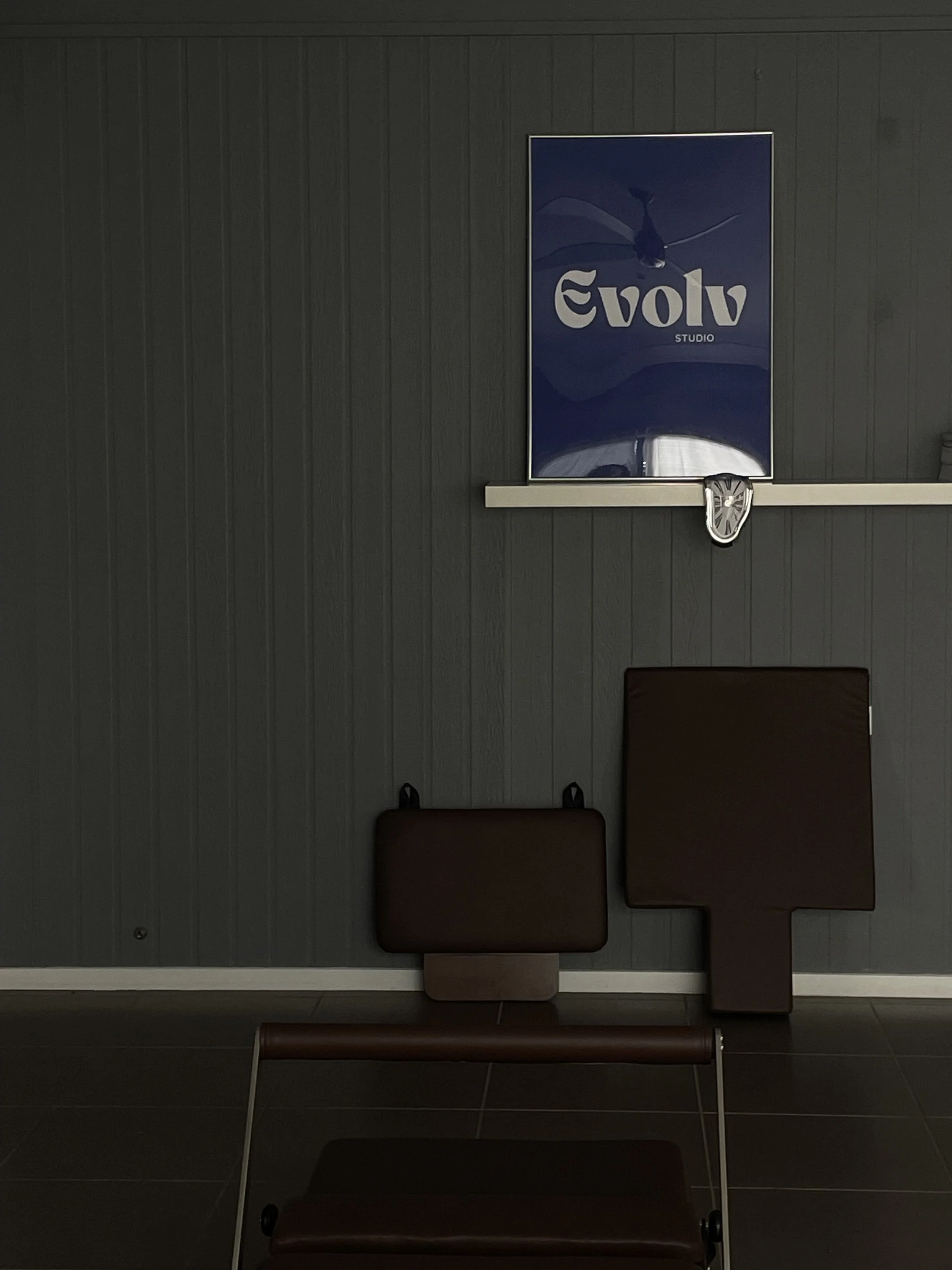 Brown reformer jump board and pregnancy wedge, and a blue Evolv Studio poster on a dark gray wall.
