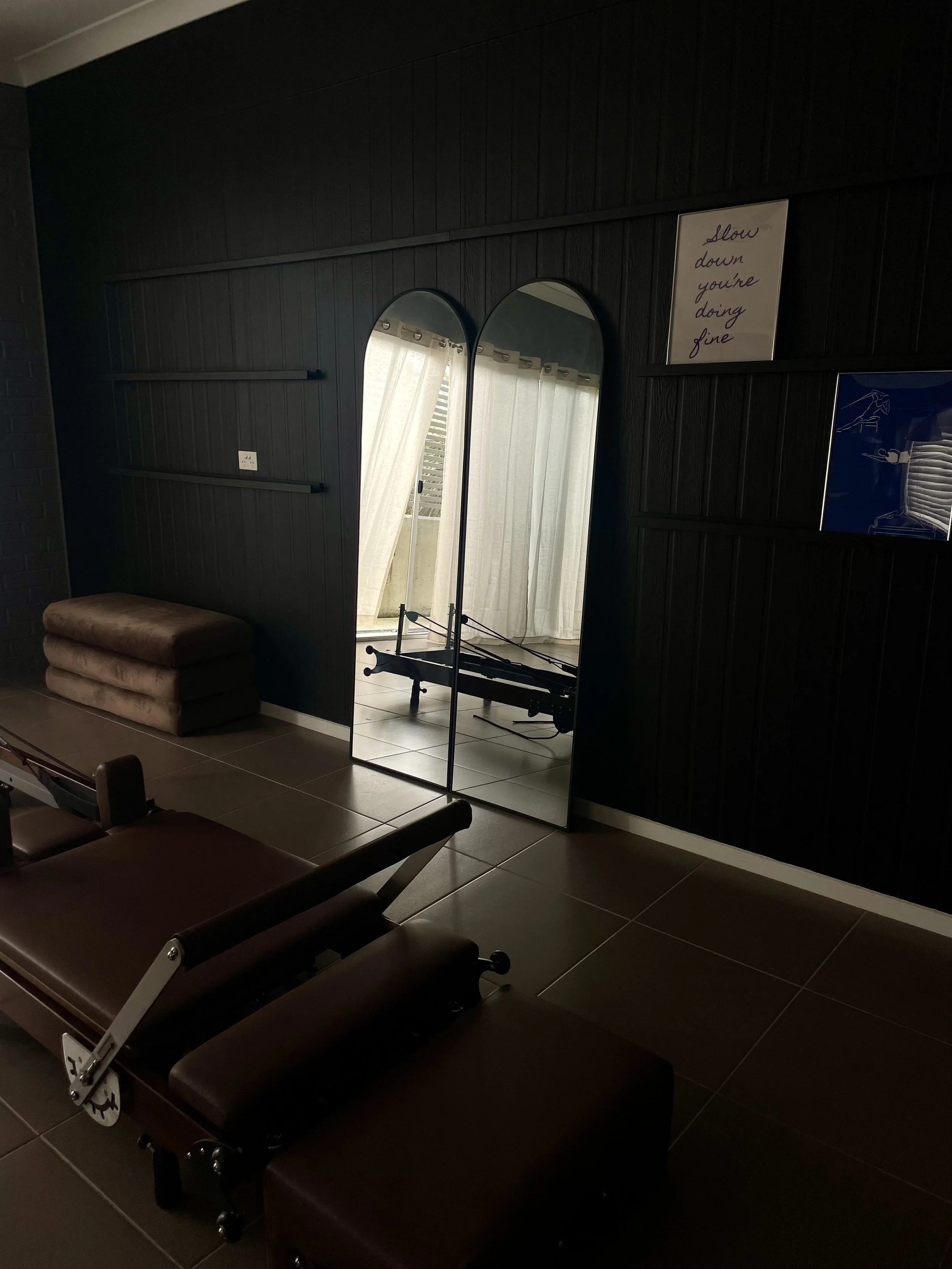 Dark interior room with amplitude black wall, round and rectangular mirrors, a painting with a motivational quote, a brown velvet bench, and a brown reformer