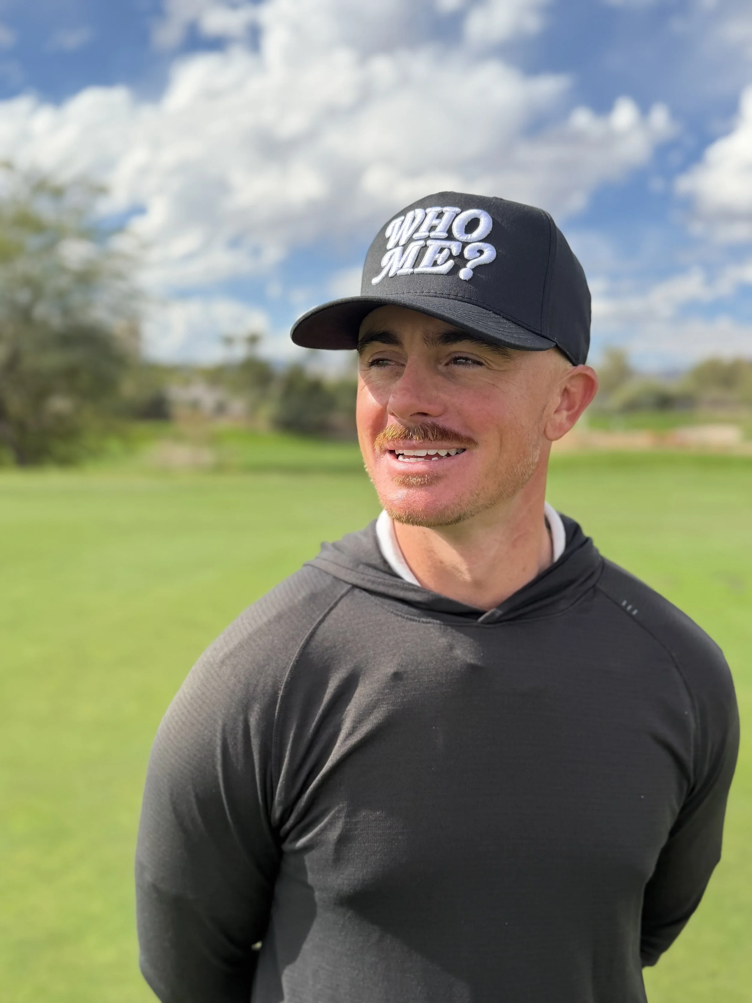 A man with a mustache wearing a black cap with the words 'WIE O ME?' and a black hoodie, standing outdoors on a grassy field with trees and a partly cloudy sky in the background.