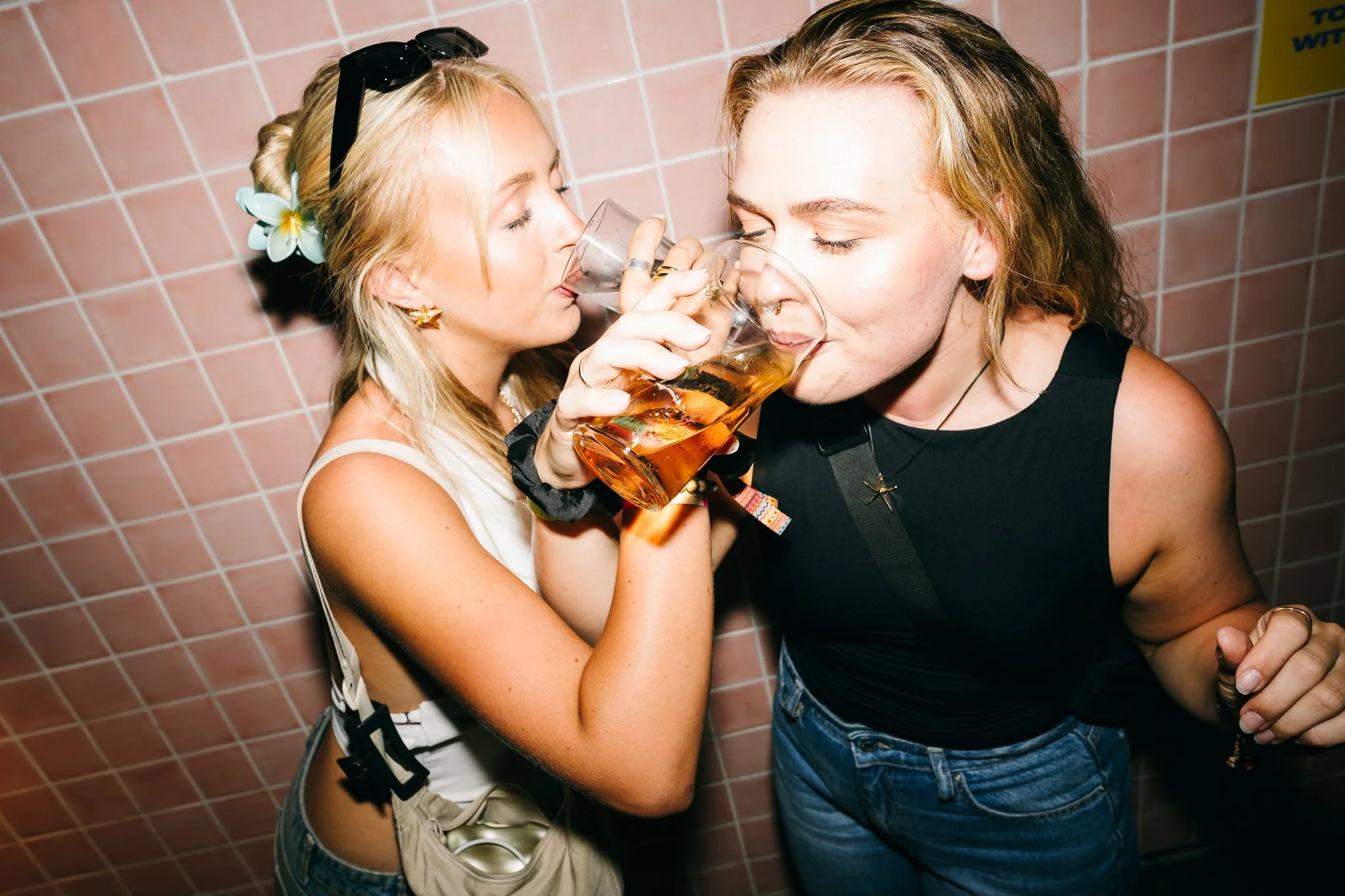 Two women with light skin drinking beer from glasses at a party, standing close to a tiled wall.