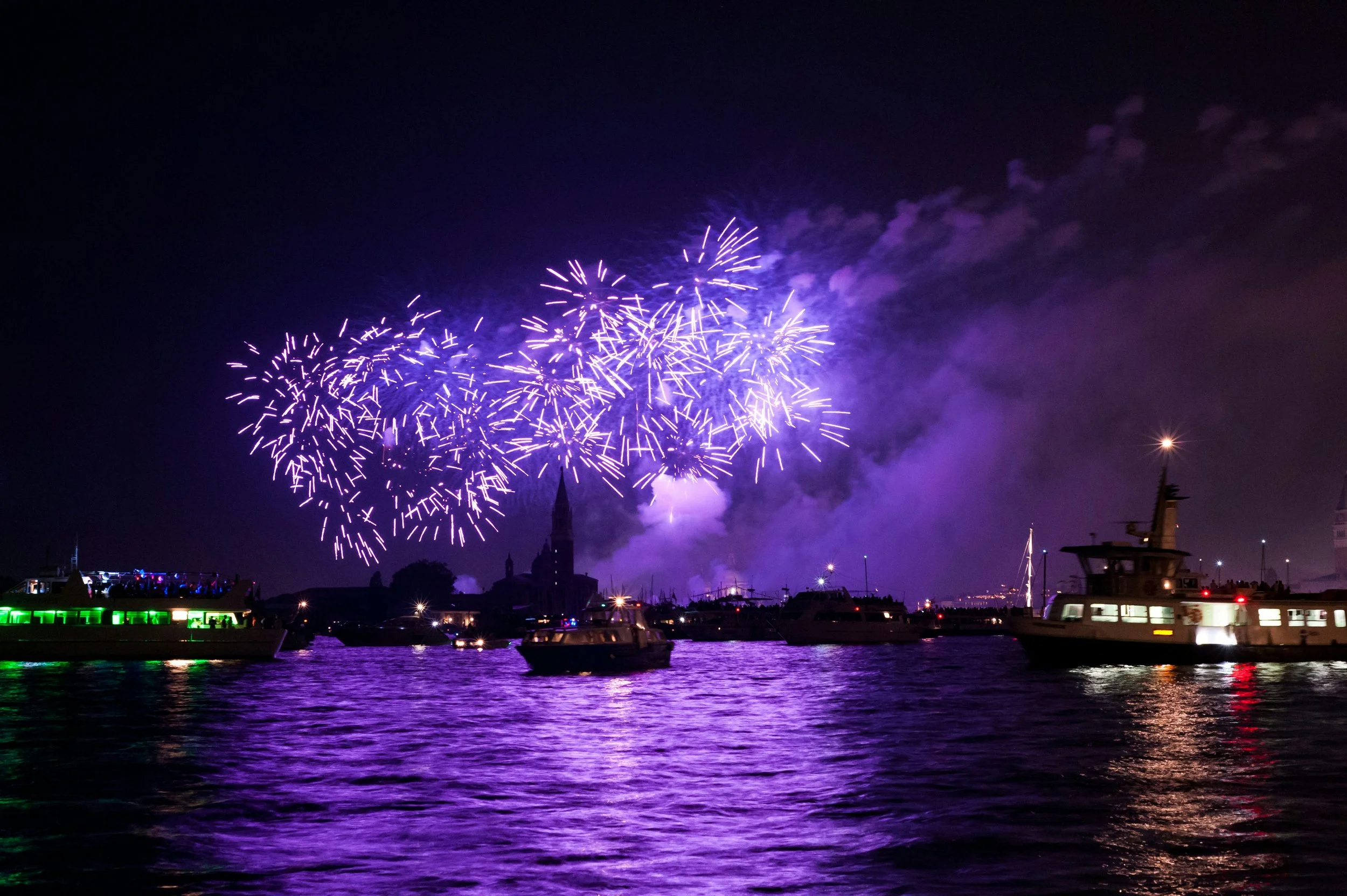Fireworks display over a water cityscape at night with boats on the water and purple smoke from fireworks.