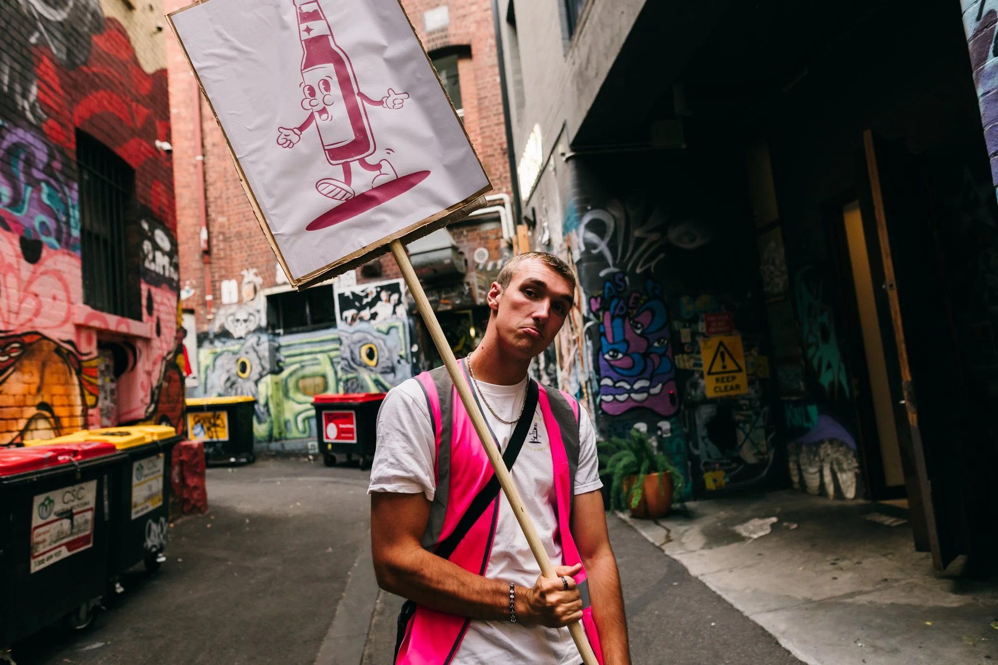 A young man standing in an alleyway with colorful graffiti, holding a sign with a cartoon soda bottle giving a thumbs-up.