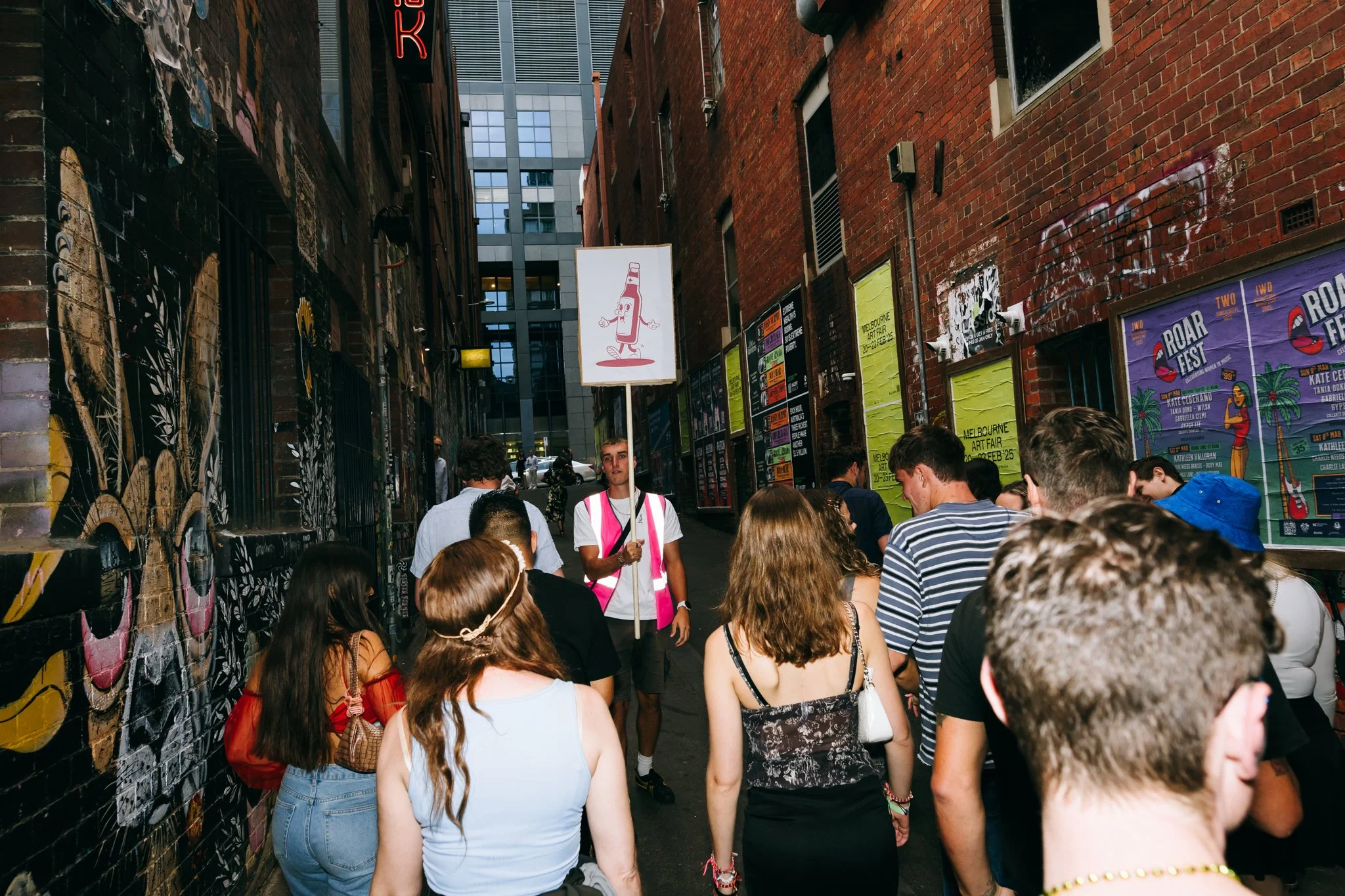 Crowd of young people walking in a narrow alleyway decorated with graffiti, posters, and colorful murals, with a man holding a sign featuring a cartoon ketchup bottle.