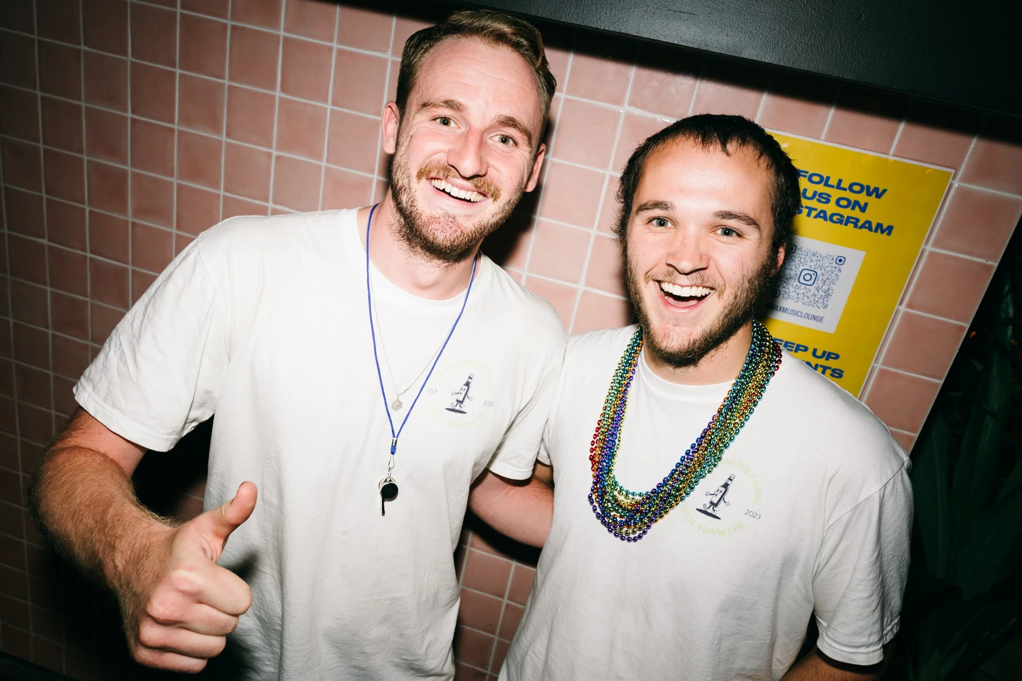 Two smiling men with light skin and beards standing close together, wearing white T-shirts with similar logos, one giving a thumbs-up, in front of a pink tiled wall and a yellow sign encouraging Instagram follow.