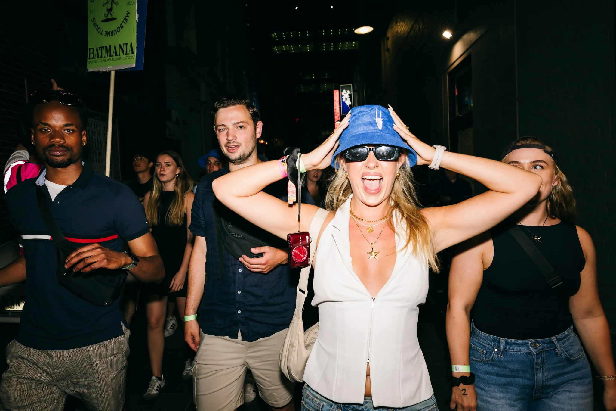 Group of young adults walking in a dark urban street at night, with one woman in the front wearing sunglasses, a blue hat, and expressing excitement.