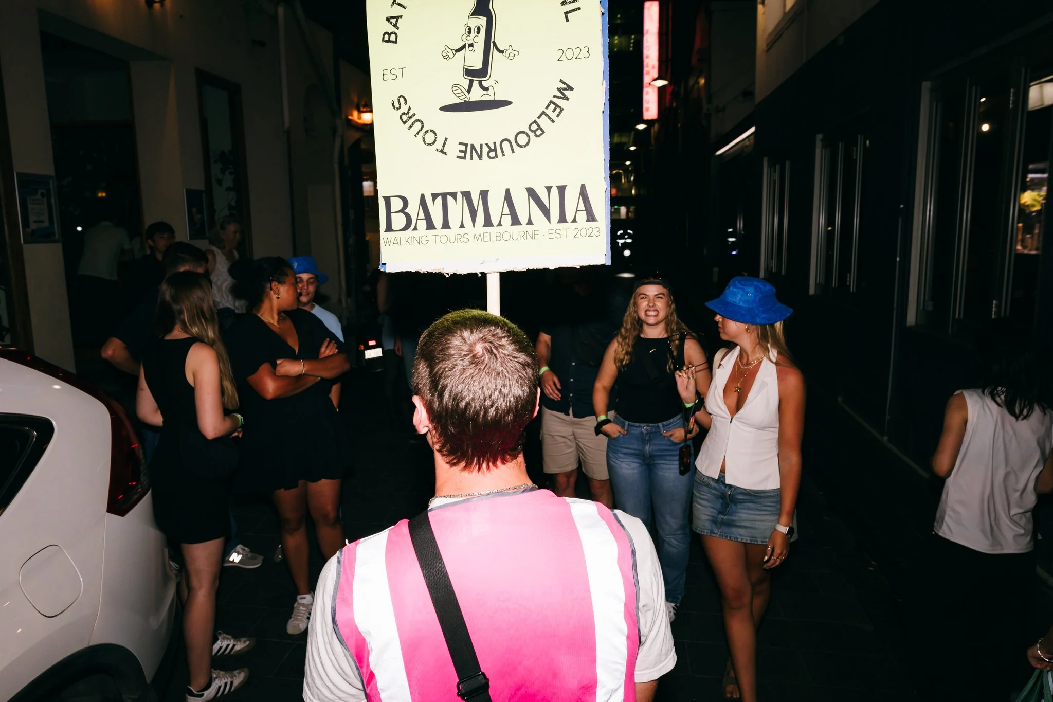 A group of people gathered around a man with his back to the camera, standing under a Batman-themed sign that reads 'BATMANIA WALKING TOURS MELBOURNE - EST 2023'. The scene appears to be on a city street at night.
