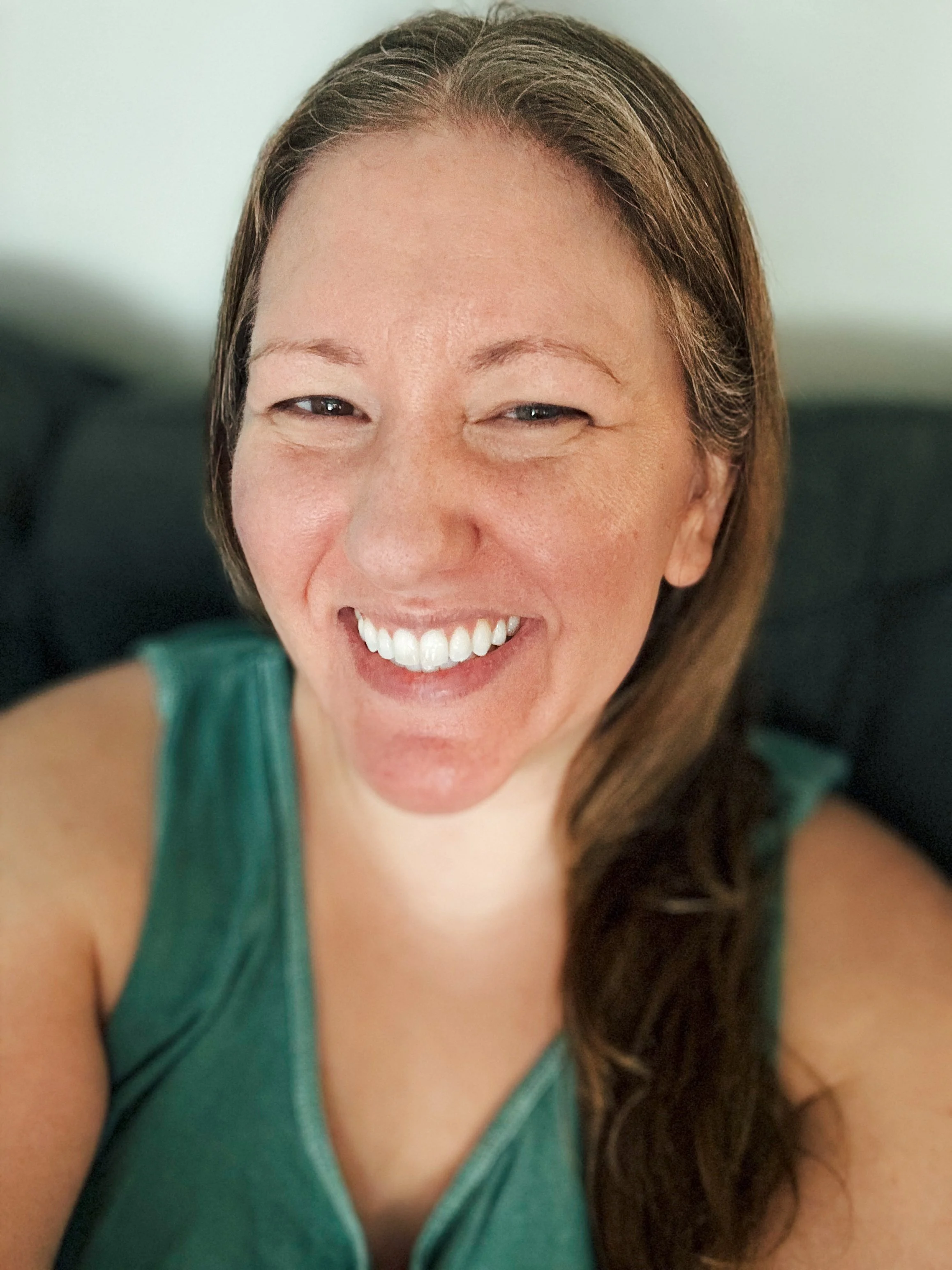 A woman with long brown hair smiling at the camera, sitting on a dark sofa, wearing a green sleeveless top.