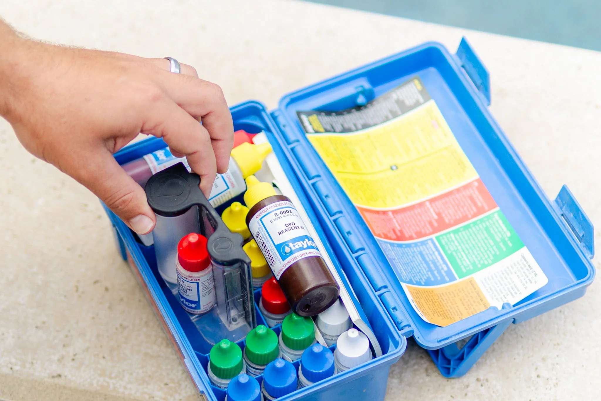 A person is reaching into an open blue water chemistry kit with various test bottles and water chemistry  supplies inside on a beige pool surface.