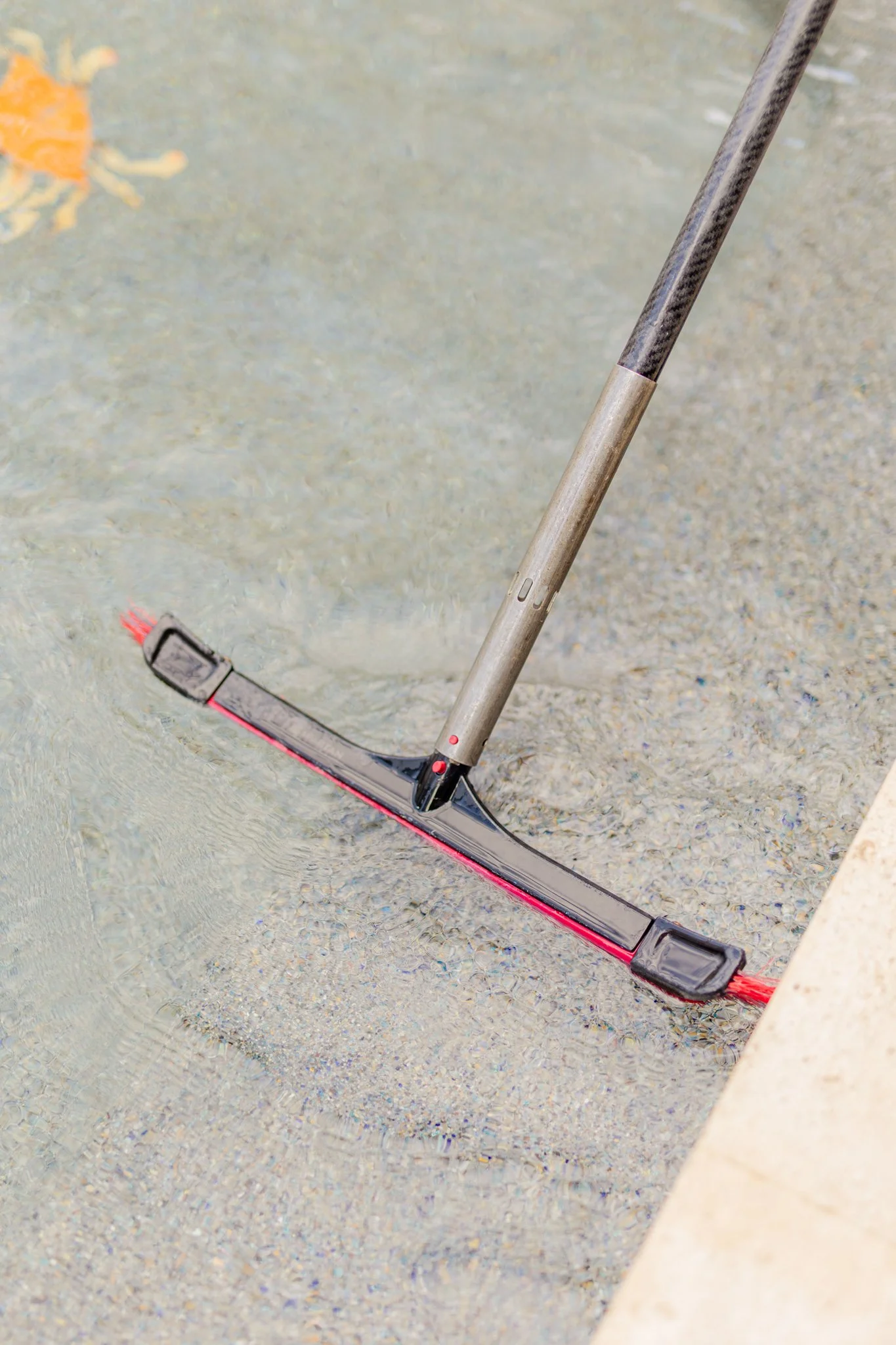Pool cleaning brush submerged in a shallow pool with water and textured flooring.