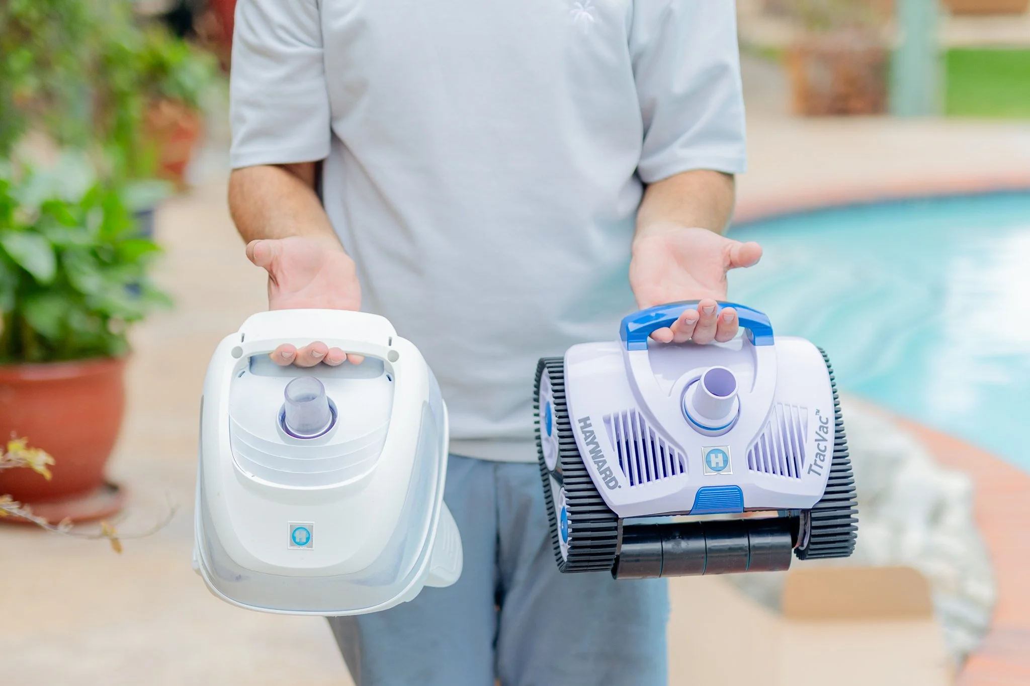 A person holding two portable pool cleaning robots, one in each hand, near a swimming pool. One in his right hand is an older broken cleaner that is being replaced by the one in his left hand which is a new Hayward TracVac