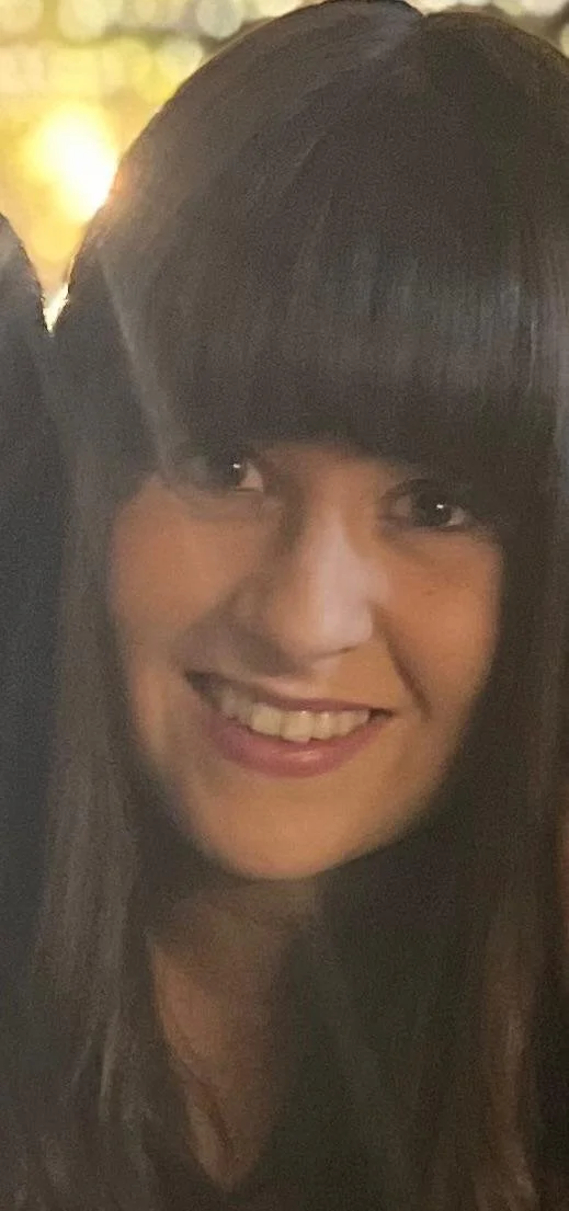 Smiling woman with long dark hair and blunt bangs in a dimly lit environment.