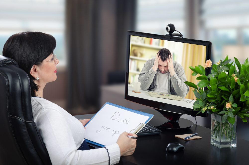 A woman sitting at a desk in an office holding a sign that says 'Don't give up' while talking to a man appearing distressed on a video call on her computer screen.