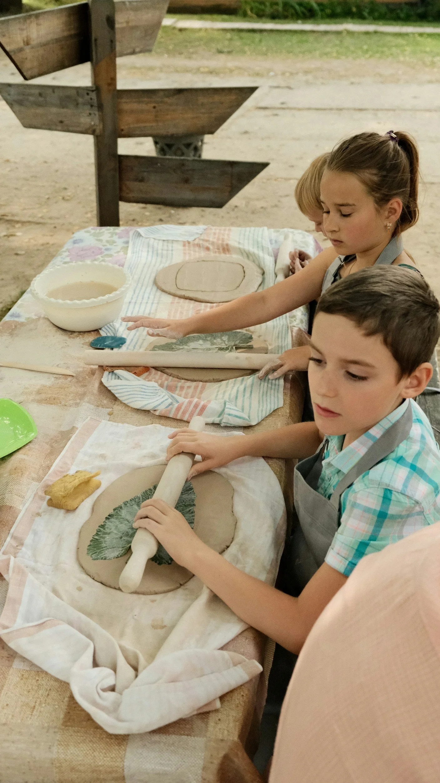 Children making clay art outdoors, with rolling pin and leaf imprints on clay.