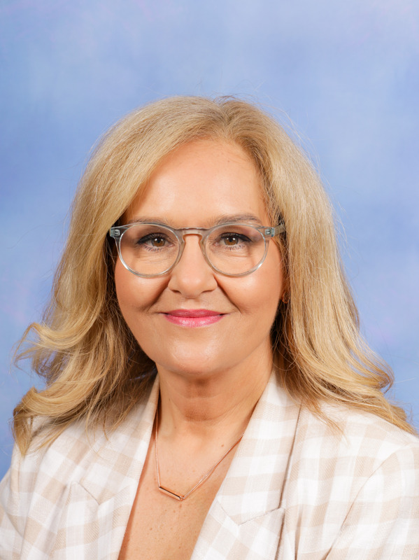 Portrait of a middle-aged woman with blonde hair, wearing glasses, a light-colored checked blazer, and a necklace, smiling against a blue background.
