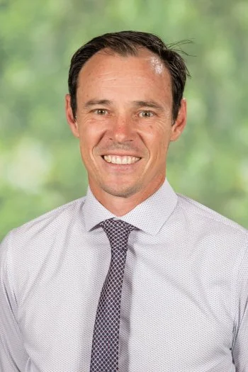 A smiling man in a white dress shirt and patterned tie, standing outdoors with a blurred green background.