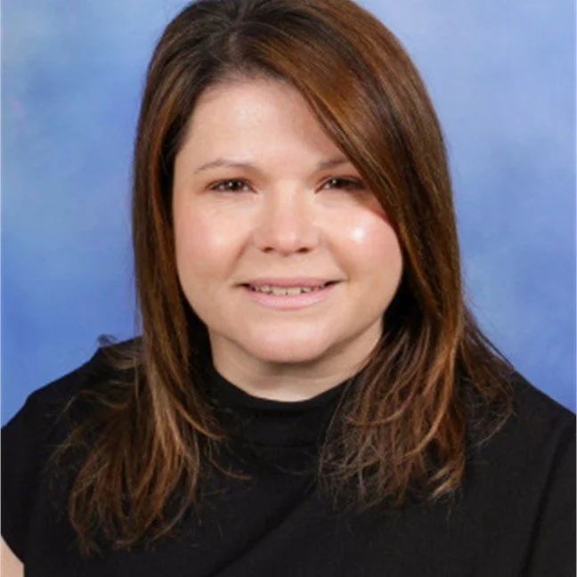 A woman with shoulder-length brown hair, wearing a black top, smiling, with a blue background.