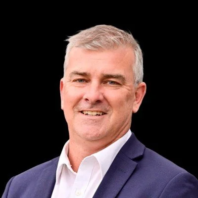Professional portrait of a middle-aged man with gray hair, smiling, wearing a dark blue blazer and white shirt, against a black background.