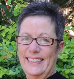 A close-up of a smiling woman with short dark hair and glasses, outdoors with green foliage in the background.