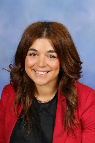 A woman with long brown hair smiling, wearing a red blazer over a black top, against a light blue background.