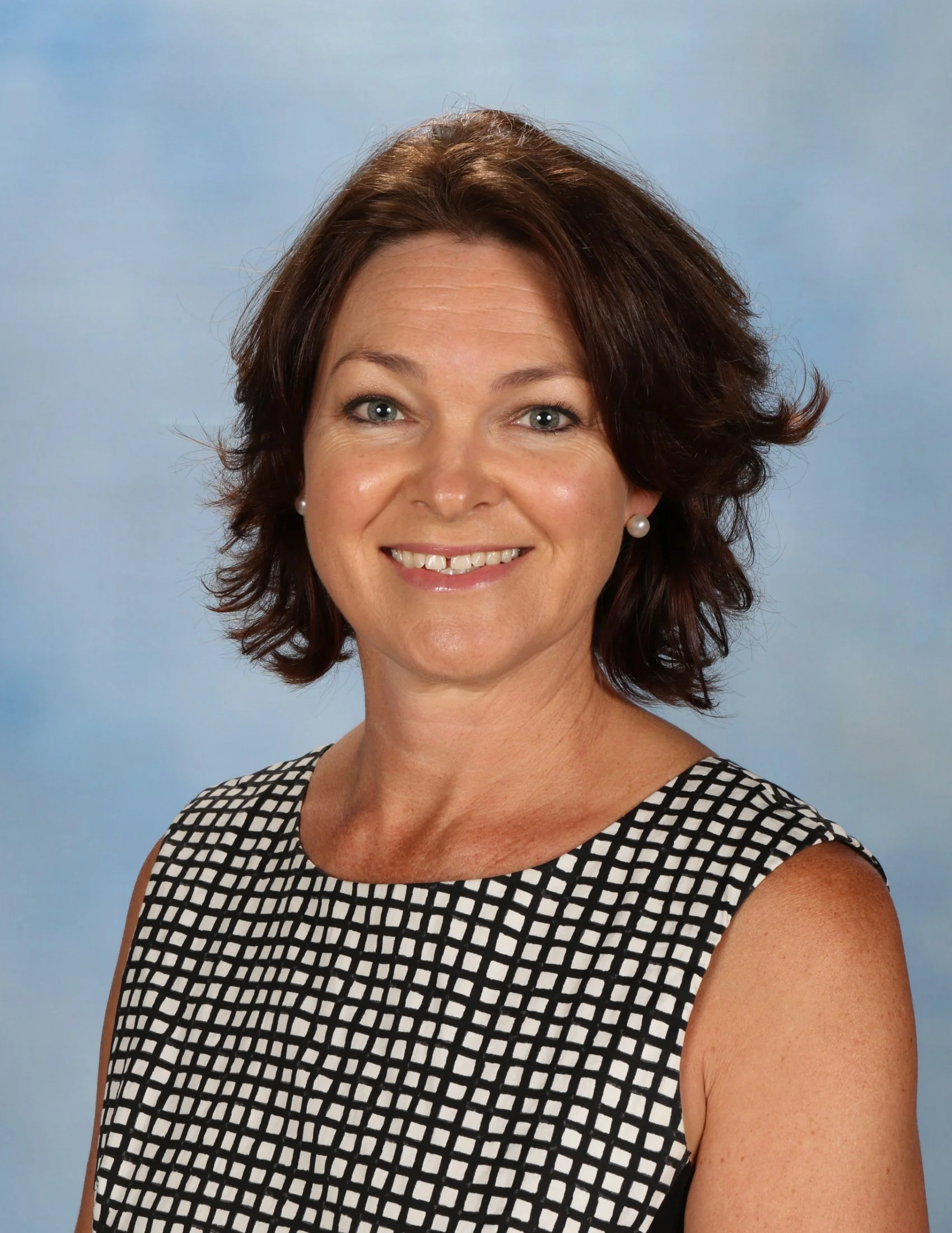 A woman with short, wavy brown hair, blue eyes, and light skin, smiling at the camera, wearing pearl earrings and a sleeveless black and white checkered top, against a light blue background.