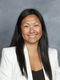 Smiling woman with long black hair wearing a white blazer and black top against a gray background.