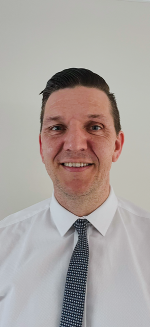 A man with dark, slicked-back hair wearing a white dress shirt and a patterned tie, smiling at the camera against a plain light-colored background.