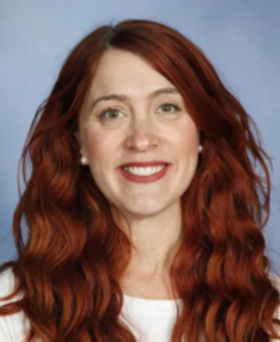 Smiling woman with long red hair, wearing pearl earrings and a white top, against a light blue background.