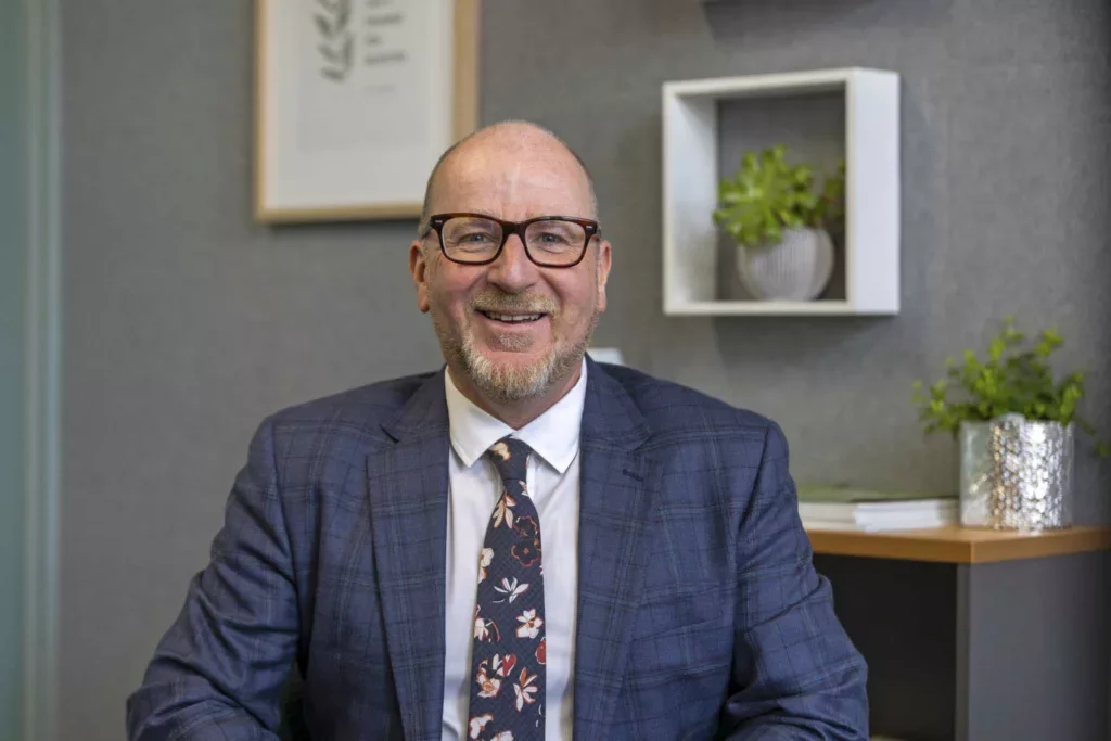 A smiling man with glasses, a beard, and a bald head, wearing a blue plaid suit and a floral tie, sitting in an office with gray walls, shelves with plants, and framed artwork.