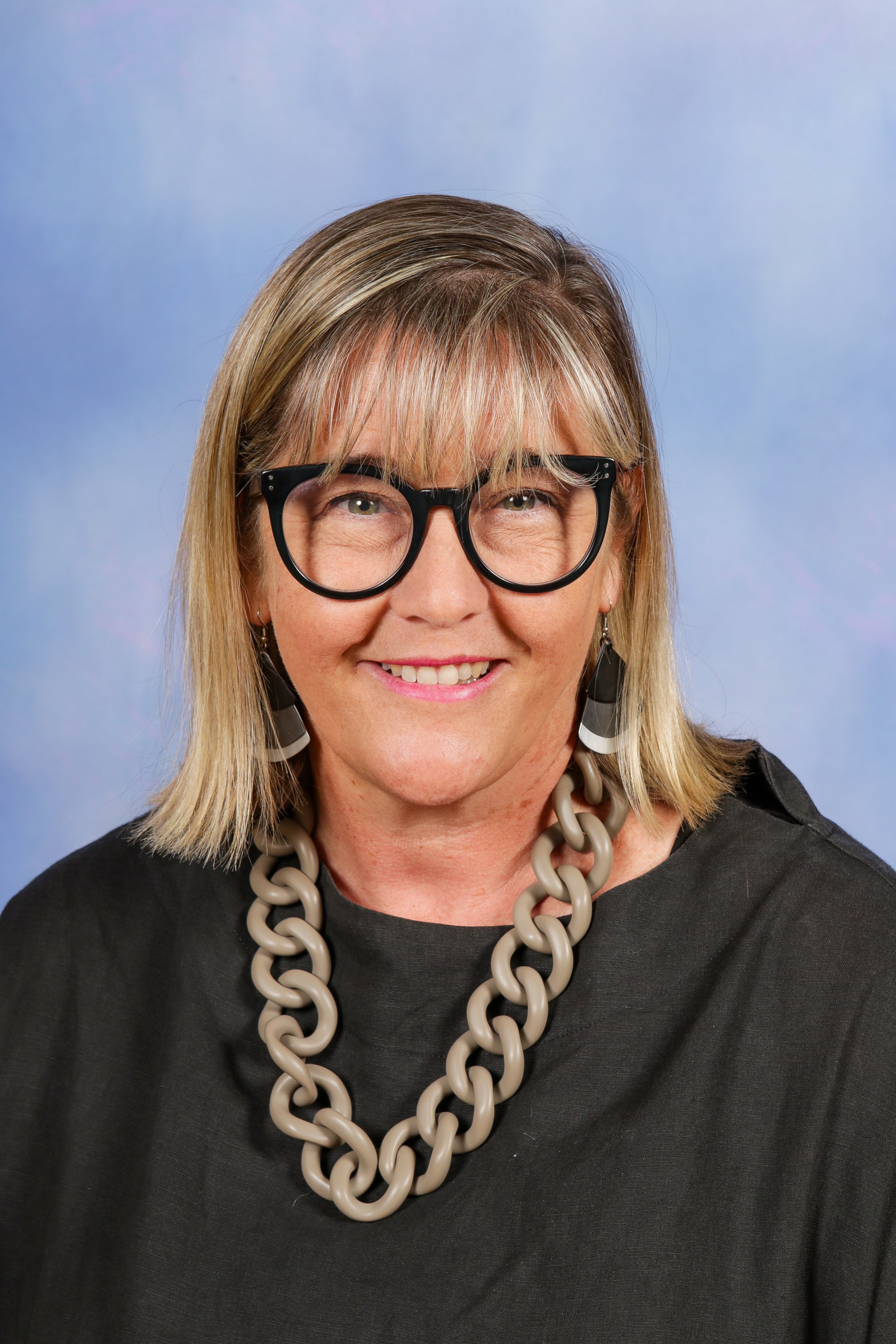 A woman with shoulder-length blonde hair, wearing black glasses, earrings, a chunky necklace, and a black top, smiling in front of a blue background.