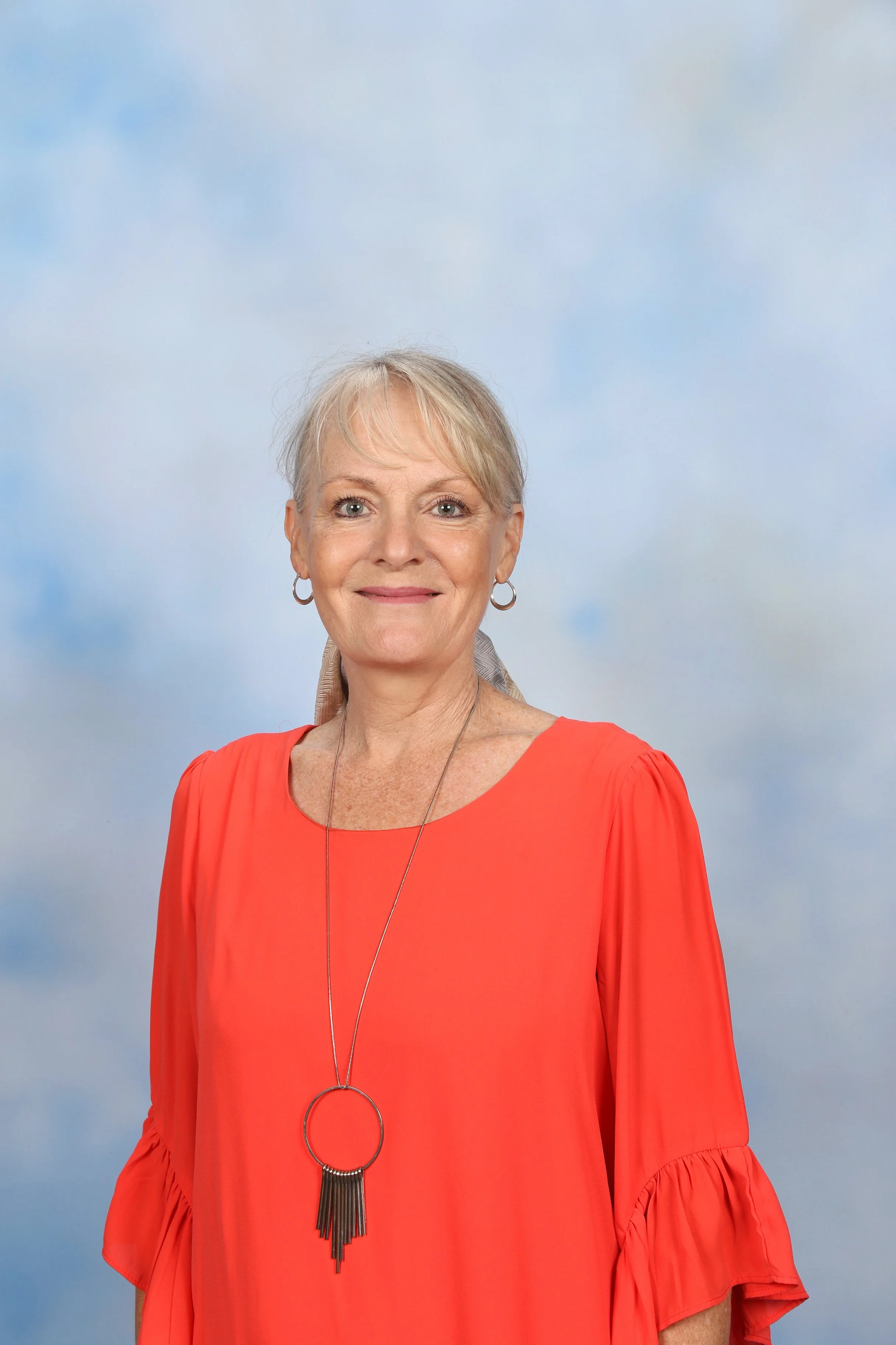 A smiling older woman with short blonde hair wearing a red dress with ruffled sleeves and a long necklace with black fringes, standing outdoors with a cloudy sky background.