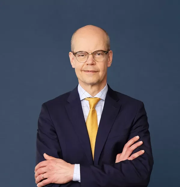Man in a dark suit, light blue shirt, and yellow tie with glasses, standing against a plain blue background.