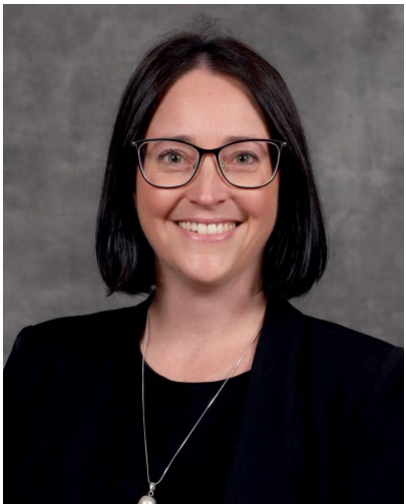 A woman with shoulder-length dark hair, wearing glasses, a black top, and a black blazer, smiling in front of a gray background.