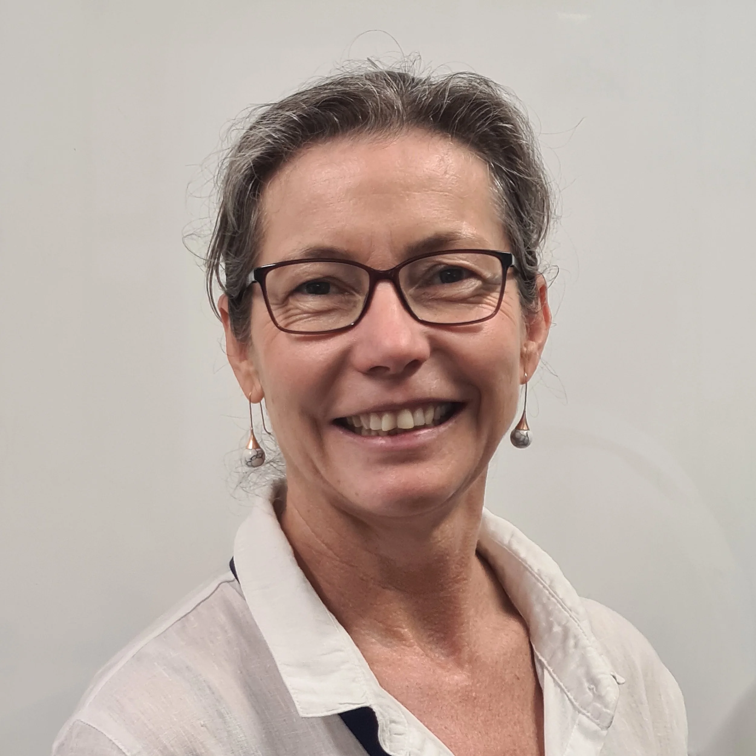 Smiling middle-aged woman with short gray hair, glasses, earrings, and a white blouse standing against a plain wall.