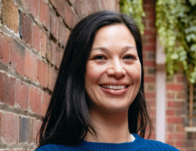A woman with black hair smiling at the camera while standing outdoors near a brick wall with greenery in the background.