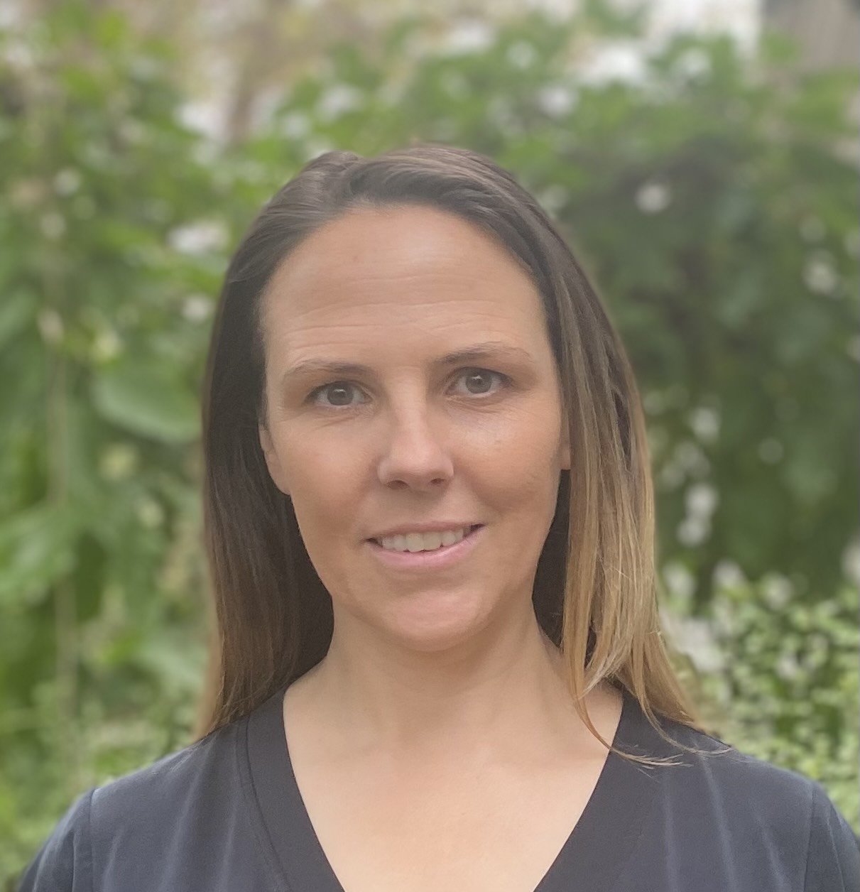 A woman with light brown hair and blue eyes smiling outdoors with green foliage in the background.