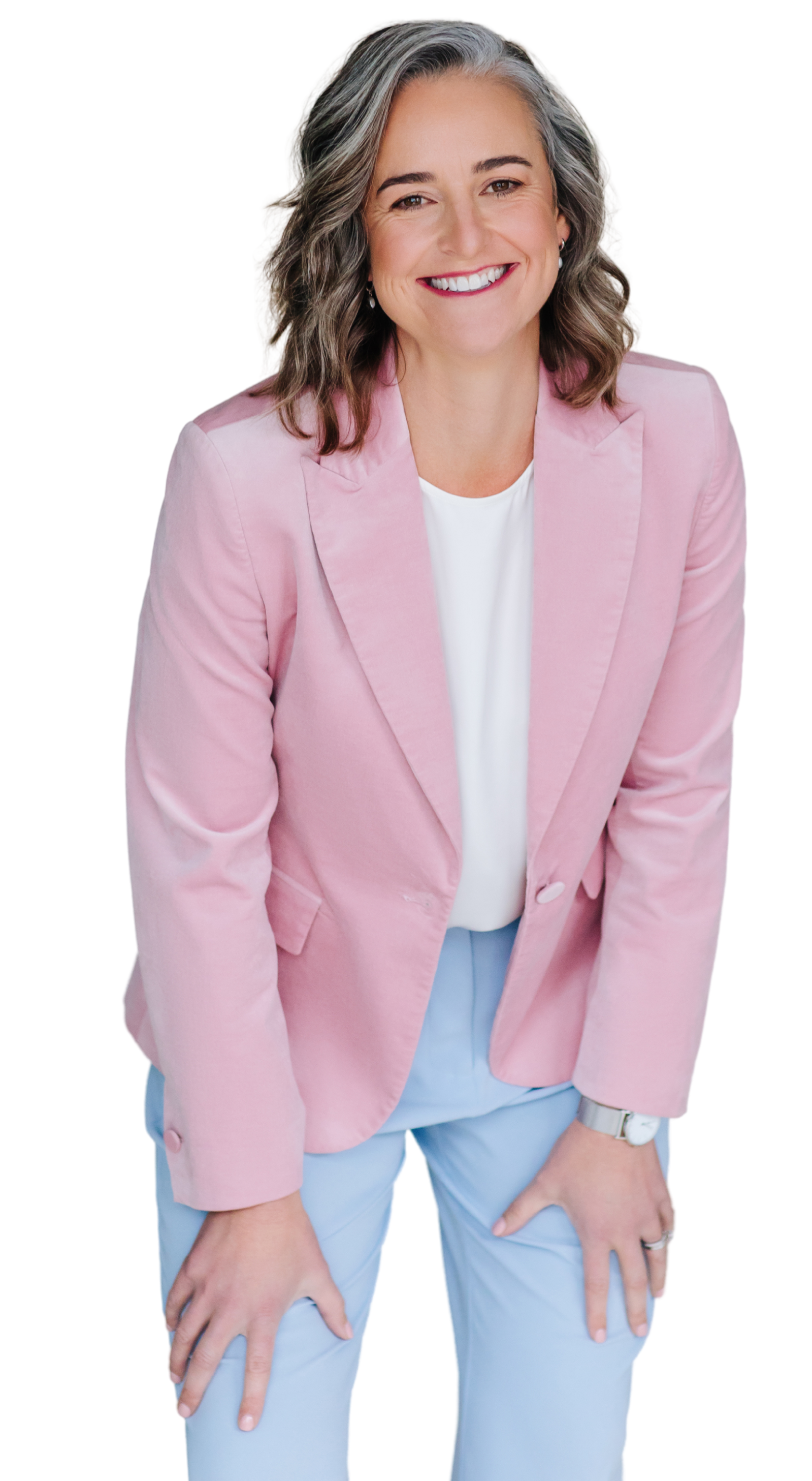 A woman with shoulder-length wavy gray hair smiling, wearing a pink blazer over a white blouse, and light blue pants.