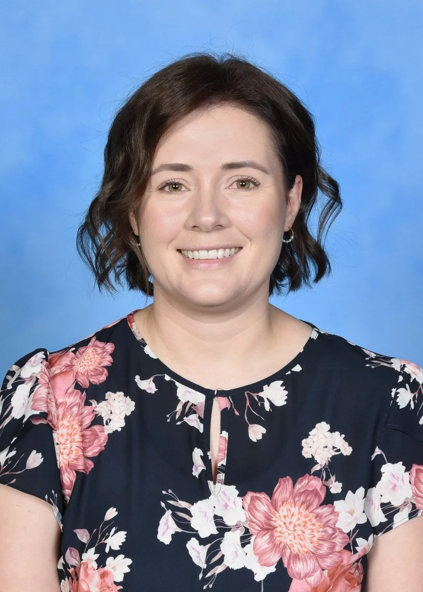 A woman with shoulder-length wavy brown hair, wearing a dark floral blouse, smiling against a blue background.