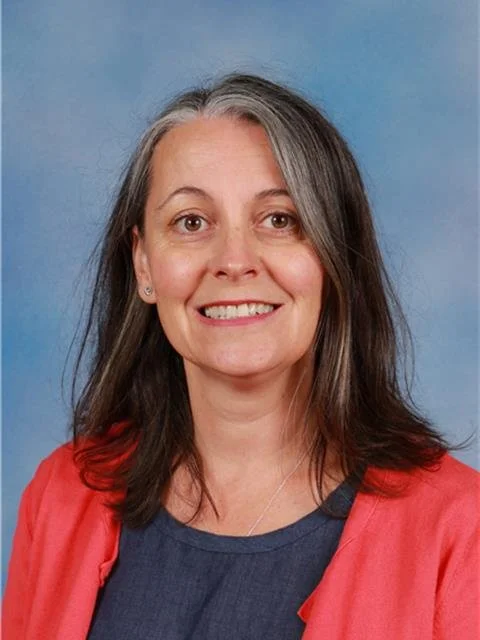 A woman with shoulder-length gray hair, wearing a dark blue top and a red blazer, smiling in front of a blue background.