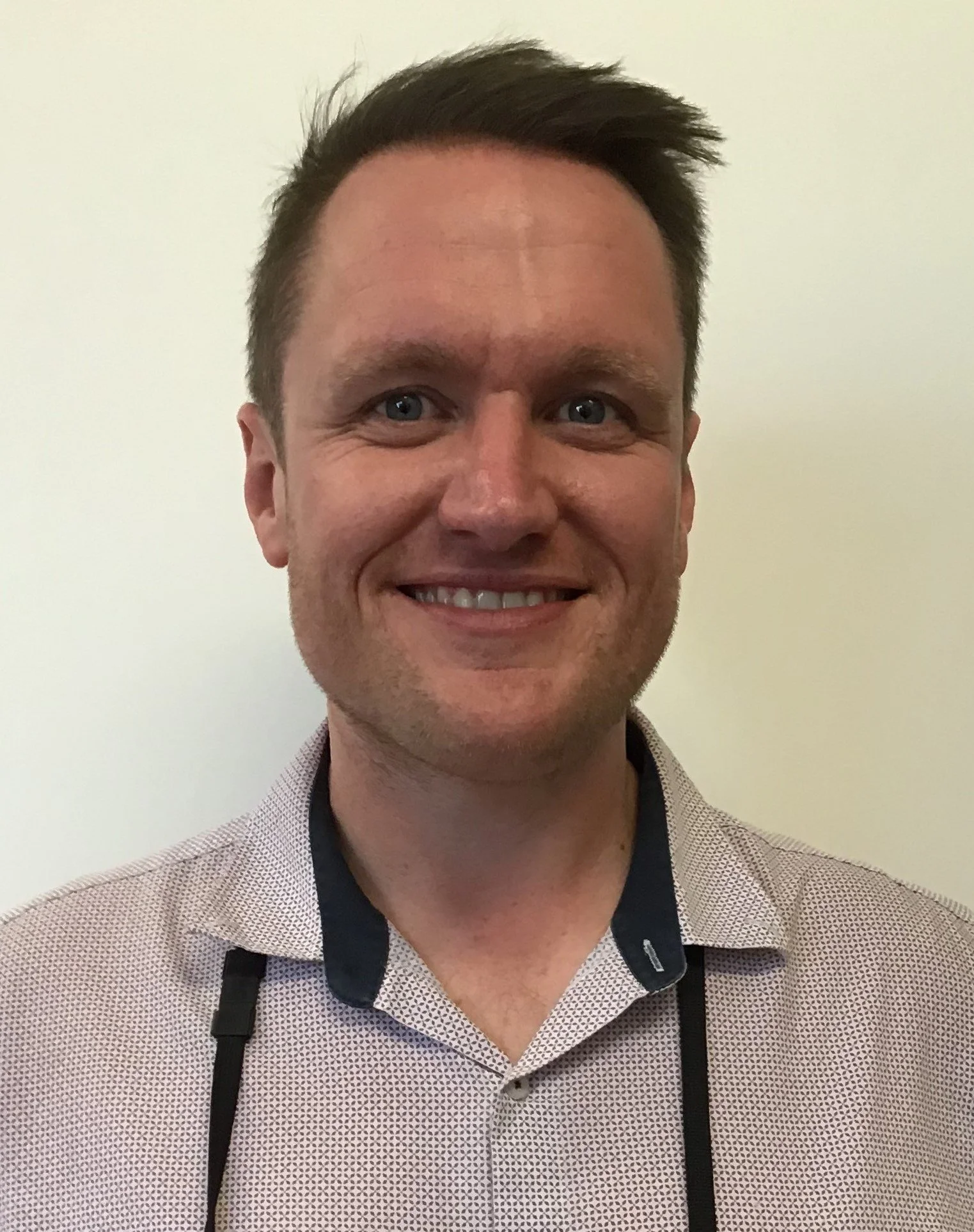 A smiling man with short brown hair, blue eyes, and a patterned shirt standing against a plain wall.