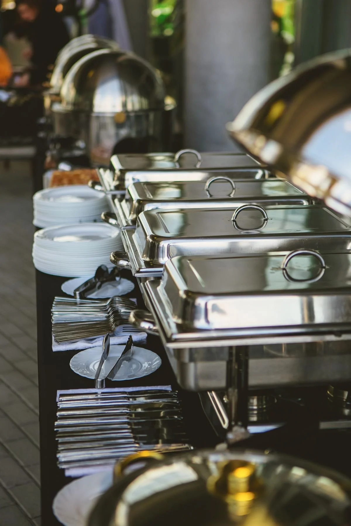 A buffet table with stainless steel chafing dishes, white plates, and silverware ready for serving food.