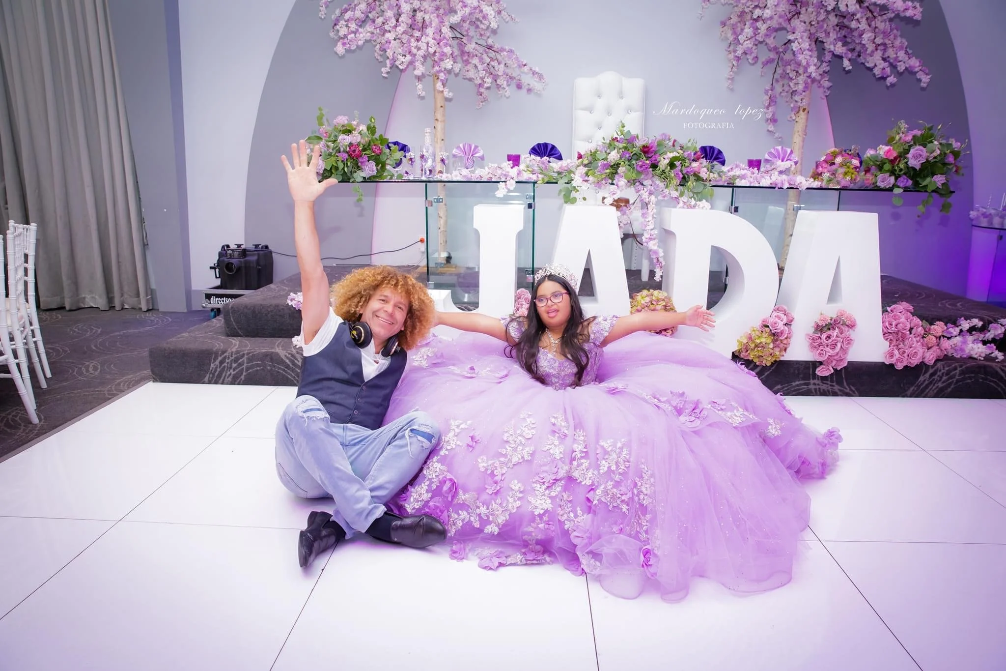 A young girl in a purple, floral-style dress with a tiara sits on a decorated stage at her Quinceañera, with a man sitting beside her. The stage features large white letters spelling “FADA,” purple and pink floral arrangements, and two flowering trees in the background.