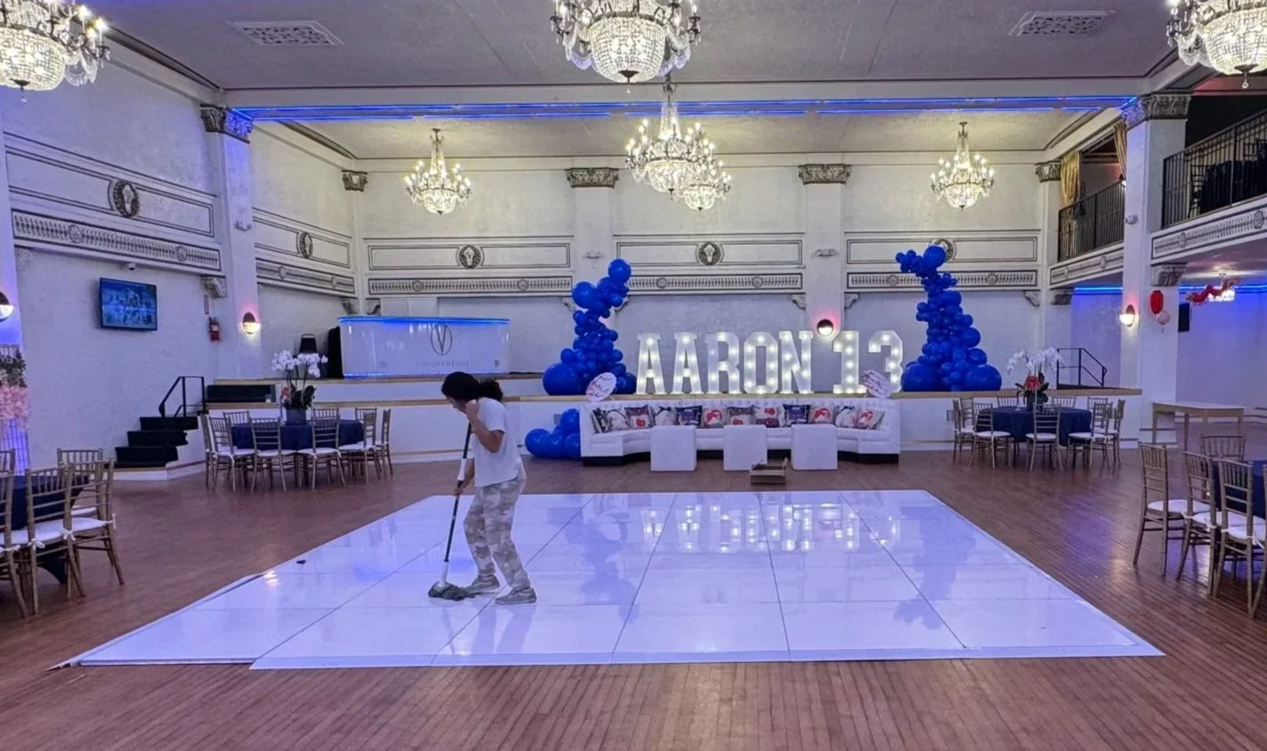A person mops the dance floor in a decorated banquet hall with balloons, large illuminated letters, and chairs set up for a celebration.